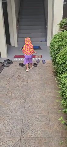 A young child wearing a pink and yellow heart-patterned headscarf and a purple dress stands facing a set of stairs. In front of the child, on a tiled walkway, are several pairs of shoes, including sandals and flip-flops. To the right of the child, a lush green bush is visible. Behind the child, a staircase leads upwards, with a blue mat and a red rug placed at its base. The text "BATAS SUCI" is visible on the red rug, which translates to "pure limit" or "cleanliness boundary" in Indonesian, suggesting a designated area for removing shoes. The child appears to be preparing to ascend the stairs or engage in an activity related to the area.