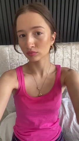 This media file is a close-up, selfie-style video of a young woman with brown hair pulled back, wearing a bright pink tank top and striped leggings. She is sitting on a bed with a tufted headboard and white bedding. The background includes a dark, ribbed wall paneling. The lighting suggests it is daytime, indoors. The woman's expression is playful as she pouts and sticks out her tongue towards the camera. She is wearing a gold necklace with a pendant. No text is visible in the media file. The location context of Welwyn Hatfield, United Kingdom, does not provide any specific visual cues within the video itself.