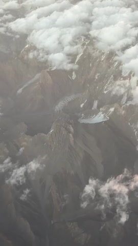 An aerial shot shows a vast mountain range from above. Clouds drift in and out, obscuring and revealing jagged peaks, steep valleys, and what appear to be glaciers snaking through the terrain. The mountains are a mix of brown and grey rock, with patches of snow visible on the higher elevations. The lighting suggests it is daytime, possibly overcast or at a time of day when the sun is low. There are no visible signs of human presence or activity.