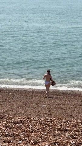 Two women in swimwear are on a pebble beach at the water's edge. One woman, wearing a light-colored bikini, walks into the water. Another woman, wearing a dark bikini, is already in the water. The sea is calm and blue, and the sky is clear.