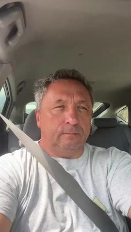 A middle-aged man with short gray hair and a gray t-shirt is sitting in the driver's seat of a car. He is wearing a seatbelt. The camera is positioned in front of him, looking directly at his face. The car's interior is visible, including the dashboard and the passenger seats. The man's expression is neutral. The video appears to be a selfie taken while driving.