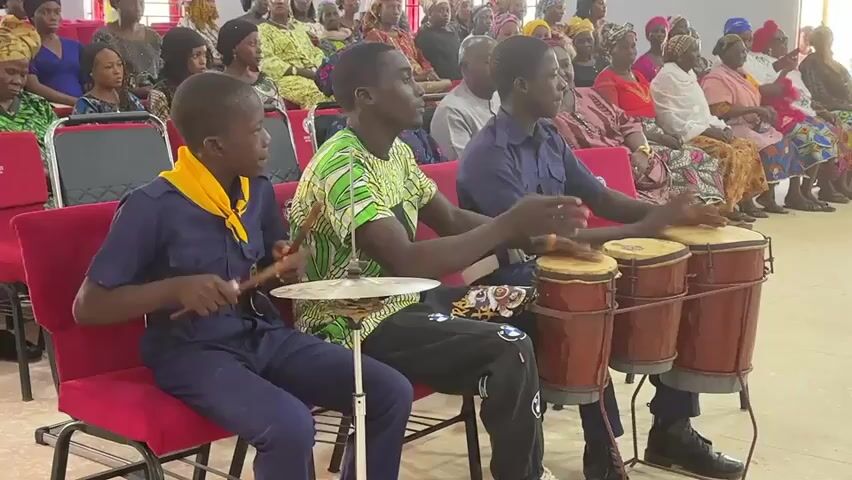 Three young men play drums in front of an audience. The audience consists of men and women of various ages. The young man on the left wears a blue uniform with a yellow scarf around his neck and plays a cymbal and drum. The young man in the middle wears a green patterned shirt and black pants and also plays a cymbal. The young man on the right wears a navy blue uniform and plays four drums. All three young men are seated.