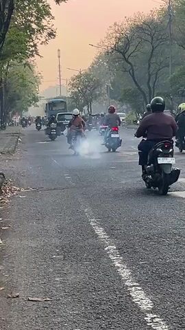 The video shows a street scene with numerous motorcycles and scooters. The vehicles are traveling in the same direction down a paved road lined with trees on both sides. The time appears to be either dawn or dusk, as the sky is a pale orange hue. Some of the motorcycles are emitting visible white smoke, likely from their exhaust. A few people are visible on the motorcycles, most wearing helmets. In the background, there is a large truck or bus, and further in the distance, there appears to be a tower or tall structure. The overall impression is of a busy street with moderate traffic.