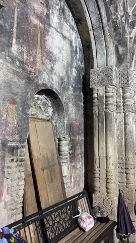 The video pans across an interior of an ancient stone building, likely a church given the religious iconography visible. The scene is dimly lit, with the primary light source appearing to be artificial, possibly from within the church. The stone architecture is aged and weathered, with visible repairs and patches on the walls and ceiling, suggesting a long history and significant preservation efforts.

The subjects in the foreground include a dark metal bench with a patterned seat. On the bench, several objects are placed: a shopping bag with some white contents, a dark umbrella, and what appears to be a patterned textile. To the left of the bench, two large wooden planks are leaning against the wall, one of which has a large green cross painted on it. The walls themselves are adorned with faded, ancient frescoes depicting figures, some with detailed faces. Ornate stone carvings are visible around doorways and columns, demonstrating intricate craftsmanship.

In the background, through an open doorway, the interior of another part of the church is visible. This area appears brighter, with more religious artifacts such as icons and possibly an altar. Several figures, likely visitors or clergy, can be seen in this background area, adding a sense of ongoing activity or worship.

Notable details include the rough texture of the stone, the peeling paint of the frescoes, and the detailed carvings that hint at a rich cultural and religious past. The overall atmosphere is one of historical reverence and solemnity, imbued with the weight of centuries. No text is clearly discernible in the video. The presence of the bench and umbrellas suggests that visitors are present, perhaps during a tour or a service. The time of day is indeterminate due to the artificial lighting.