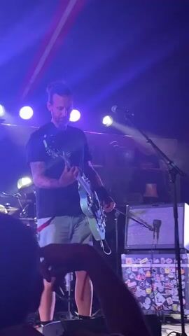 The video shows a live music performance. In the foreground, a person's arm is visible, holding up a hand with fingers spread, suggesting they are in a crowd watching the stage. The stage is lit with blue and purple lights. On the left, a drummer can be seen behind a drum kit. In the center, a man is playing a light blue bass guitar and singing into a microphone. He is wearing a black t-shirt, light-colored shorts, and white high-top sneakers. Behind him, there are amplifiers and other stage equipment covered in stickers. To the right, another man is playing a black bass guitar, wearing a black t-shirt, dark shorts, a cap, and checkered socks. He is also looking towards the audience. The overall environment appears to be an indoor venue with a tent-like ceiling structure visible in the background.