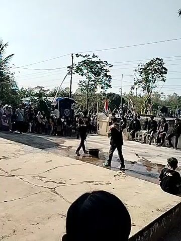 A group of people are gathered outdoors, seemingly for a performance or event. In the foreground, a person sits facing away from the camera. In the midground, two individuals dressed in black are performing an action that appears to involve rhythmic movement or dance. One individual is holding what looks like a stick or pole. They are performing on a wet, paved surface with a bucket nearby. A crowd of spectators is gathered in the background, some seated and some standing, watching the performance. There are trees, buildings, and utility poles visible in the environment. Indonesian flags are displayed, suggesting the location might be Indonesia. The sky is clear and bright, indicating it is daytime.