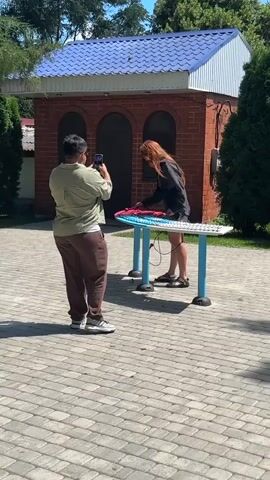 In the video, a person with dark hair and a green shirt stands holding a phone in landscape mode, filming a person with long red hair who is standing on a table. The person on the table is wearing a black jacket and shorts and appears to be interacting with a colorful, flat object on the table. The table has blue legs and is situated outdoors on a paved surface. Behind the table and the people is a brick building with a blue roof and three arched doorways. Trees and greenery are visible in the background. The sun appears to be shining, casting shadows on the ground. The overall scene suggests an outdoor activity or performance being documented.