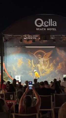 The video shows an outdoor performance at night. In the background, a stage is set up with a large backdrop featuring a painting of an angel with wings spread. Above the backdrop, a black awning displays the logo "Qella Beach Hotel". On the stage, an individual wearing an orange shirt and white helmet is performing on a red ramp-like structure. The performer is in mid-air, appearing to be launching from or landing on the ramp. There is a bright spotlight on the performer. In front of the stage, an audience is seated in chairs, facing the performance. Many audience members are holding up their phones to record the show. The environment appears to be an outdoor seating area, possibly a resort or hotel venue, with tables and chairs visible. The lighting is dim, consistent with nighttime.