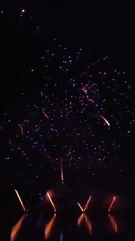 A fireworks display is happening at night. The fireworks are in the shape of an X and are reflecting on the water. There are also some sparks in the sky from the fireworks.