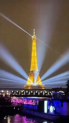 The video shows the Eiffel Tower illuminated with bright yellow lights and surrounded by white spotlights emanating from the base and reaching upwards into the night sky. In the foreground, a boat with glowing purple and blue lights is partially visible on a body of water. The scene is dark, indicating nighttime.
