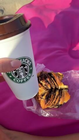 A hand holds a white Starbucks coffee cup with a brown lid. Next to the cup, on a bright pink fabric, rests a partially eaten pastry, wrapped in clear plastic. The background shows a sandy beach and the pink fabric. The video is 5 seconds long.