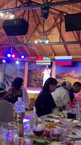 A performer in a white suit is singing on a stage. Two flags are hanging behind the performer, one appears to be the flag of Abkhazia and the other the flag of Russia. The stage is set against a backdrop of mountains. In the foreground, several people are seated at tables, with food and drinks laid out. The environment appears to be an indoor venue with a wooden ceiling and exposed beams. Lighting from stage lights illuminates the scene.