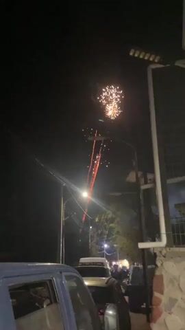 At night, fireworks light up the sky with light streaks and sparkles above a residential neighborhood street. The camera looks upwards at the fireworks, with foreground objects including an overhanging street light with a bright glow, power lines, cars, a section of a building, and a fence.  Silhouettes of people can be seen further down the street.