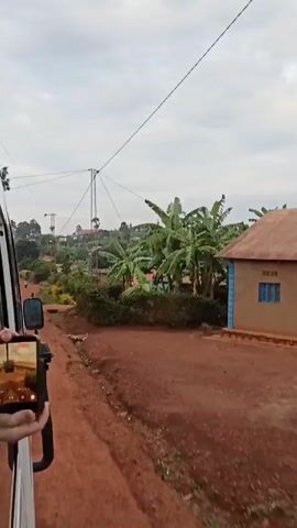 The video shows a red dirt road lined with greenery. A building with a red roof and banana trees stand on the right. A telephone pole stretches toward the sky.  A person stands on the side of the road, looking toward the camera. A person inside a vehicle films the scene on a cell phone.