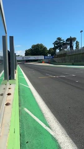 The video shows two race cars speeding along a racetrack. The track is bordered by a green wall and a safety barrier. The sky is clear and blue, and the lighting indicates it is daytime. A sign spanning across the track displays the words “REGIONE LAZIO” on one side and “Sparco” on the other. The cars are a vibrant red and a sleek silver. The red car races by first followed by the silver car. The cars are the main focus of the video.