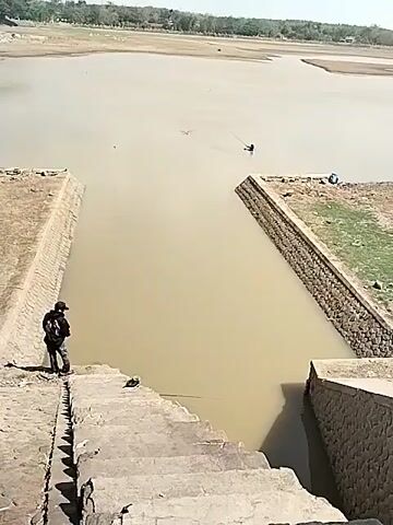 The video shows a person standing on a concrete staircase leading down to a body of muddy water. The person is wearing a dark jacket and a backpack. The water appears to be very shallow in places and has a brown, murky color. There are concrete structures on either side of the staircase, forming a channel into the water. In the distance, two other people can be seen in the water, one appearing to be fishing. The surroundings are dry and barren, with some sparse vegetation visible in the background. The sky is clear and the lighting suggests it is daytime.
