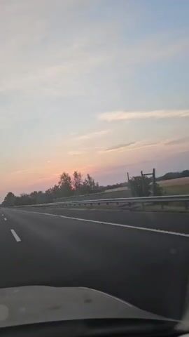 The video shows a view from inside a car driving on a highway. The sun is setting, casting a warm glow on the sky and trees on the side of the road. The highway has multiple lanes and a guardrail separating the lanes from the opposite direction of traffic. The sky is a mix of blue and pink, with some clouds. The trees appear to be deciduous and are in full leaf. The road is asphalt and appears to be in good condition. The video is taken during daylight hours, likely late afternoon or early evening.