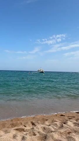 The video shows a beach with a boat on the water and a few people in the distance. The sky is blue with some clouds. The sand on the beach is light brown. The water is a clear blue-green.