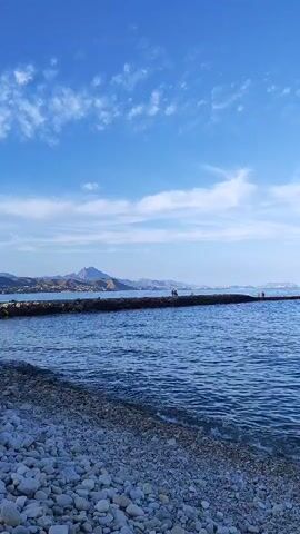 The video shows a serene seascape under a bright blue sky with scattered white clouds. In the foreground, a pebble beach slopes gently towards the calm, blue water. A stone breakwater extends horizontally across the middle ground, with several small figures walking along its length. In the background, a hazy coastline with hills can be seen. The overall impression is one of tranquility and a pleasant day at the seaside.