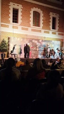 A stage performance is underway outdoors in front of a brick building with arched windows. The stage is covered with a backdrop of lights and a logo that reads "VolArtico". Actors dressed in costumes are performing, including a person in a suit, a toy soldier, and several people dressed as mice. An audience sits in chairs in the foreground, observing the performance. Some audience members are using their phones. A Christmas tree stands to the left of the stage.