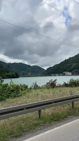 The video captures a scenic view of a large body of water, possibly a reservoir, flanked by lush green hills. In the distance, a dam structure is visible, with buildings and industrial elements suggesting a hydroelectric power plant. The sky is overcast with dramatic clouds. In the foreground, a road runs alongside the water, with a concrete barrier separating it from the vegetation. The camera appears to be mounted on a moving vehicle, indicating a journey along the roadside. No people or animals are discernible in the frame.