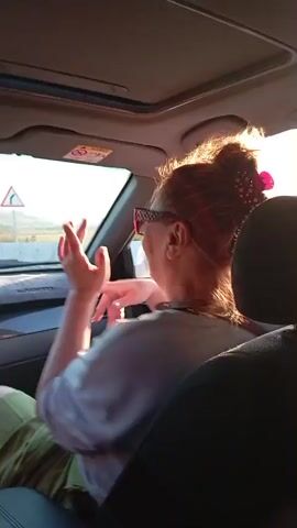The video shows a woman from the shoulders up in the passenger seat of a car. She has her hair pulled up in a bun with a pink clip. She is wearing sunglasses and a light-colored t-shirt. Her right hand is raised and she appears to be dancing or moving her fingers to music. The sun is shining brightly through the window behind her, creating a lens flare effect. The interior of the car is visible, including the dashboard and the back of the driver's seat. The video is shaky, suggesting it was filmed while the car was in motion.