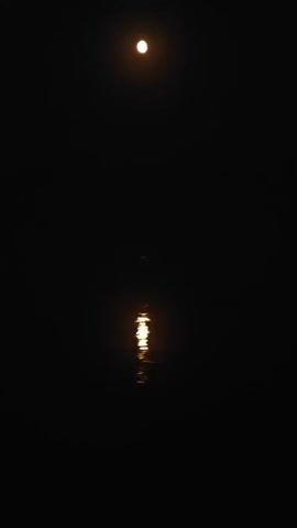 A video shows a full moon reflected in dark water. The moon is visible as a bright circle in the upper center of the frame. Its reflection appears as a shimmering, elongated band of light in the lower center of the frame. The rest of the frame is black, indicating a nighttime scene. There are no people or other objects visible.