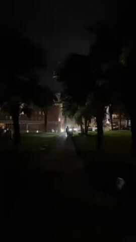 The video shows a dark outdoor scene at night. A person walks along a lit path that leads towards a building in the background. The path is flanked by palm trees. There appear to be parked cars near the building. The sky is dark with some clouds. The scene has a low light level, creating silhouettes of the trees and the person.