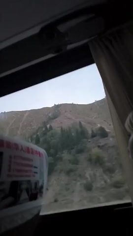 The video shows a view from inside a vehicle, likely a bus, looking out a window. The window frame and part of a curtain are visible in the foreground. A blurred object with text in a foreign language is also in the foreground, partially obscuring the view outside. Through the window, a mountainside is visible, with sparse trees and vegetation. The sky appears overcast or hazy. The overall scene suggests a journey through a mountainous, possibly rural or undeveloped area. The time stamp indicates the video lasts for approximately 4 seconds.
