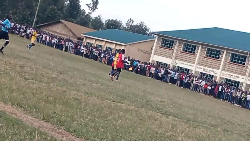 A soccer game is underway on a grassy field. There are many spectators standing on the sidelines, lining the field in front of buildings. A referee in a blue shirt and black shorts is on the field, as are several players in red and yellow uniforms. The field appears to be uneven and has a dirt path running along the bottom left of the frame. The buildings in the background appear to be educational facilities. The sky is overcast.