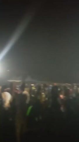 The video shows a blurry view of a crowd of people at night. Bright lights shine into the camera, making it difficult to discern details.  Tents can be seen in the background, suggesting an outdoor event or festival. The people appear to be moving and possibly dancing. The overall impression is a lively and energetic atmosphere, despite the low visibility.