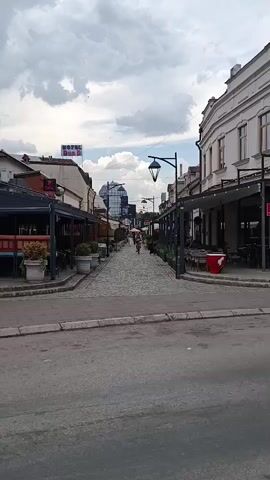 The media file depicts a street scene in Niš, Serbia, on a cloudy day. The street is paved with cobblestones and lined with buildings on both sides, many of which appear to be restaurants or cafes with outdoor seating areas. There are several people visible, including a person riding a bicycle down the cobblestone street and others walking in the distance. A blue car drives past in the foreground. A sign for "HOTEL DVA R" is visible on one of the buildings. The overall mood of the scene is calm and leisurely, with the overcast sky suggesting it might be early morning or late afternoon. The architecture of the buildings, particularly the older style on the right, hints at a historic urban setting.