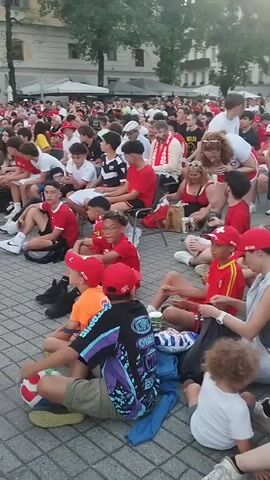 The video shows a crowd of people sitting and standing in an outdoor square, likely watching a public viewing event for a sports match. Many individuals are wearing red, and some are wearing red hats, suggesting support for a particular team. A large Swiss flag is visible in the background, along with buildings and trees, indicating an urban setting. Some people are wearing soccer jerseys, including one person wearing a German soccer jersey. The atmosphere appears to be relaxed and casual, with people sitting on the ground and interacting with each other. The lighting suggests it is daytime, possibly late afternoon or early evening.