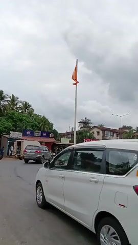 The video shows a busy traffic circle in what appears to be a tropical or semi-tropical locale. In the foreground, a white Suzuki Ertiga, identified by its yellow license plate reading "GA11T2748", is pulling into the roundabout. Multiple mopeds are circulating in the roundabout, with riders wearing helmets. The weather is overcast. In the background, there are various commercial buildings, some obscured by trees. A prominent orange flag waves atop a tall white pole in the center of the roundabout.