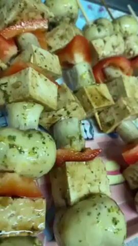 The video shows a tray of vegetarian skewers, with tofu, mushrooms, and slices of red pepper or possibly strawberries. The skewer items are coated in a green sauce. The tray is multicolored, with a design of cartoon-like characters, possibly rabbits and a hat. The camera moves slightly over the skewers, providing a close-up view.
