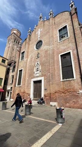 The video shows the exterior of a church in Venice on a sunny day. The church is made of brick and has a tower to the left. There are people sitting on the steps of the church, and a woman is walking in front of the building. There are three trash cans in the foreground. The sky is blue with a few clouds.