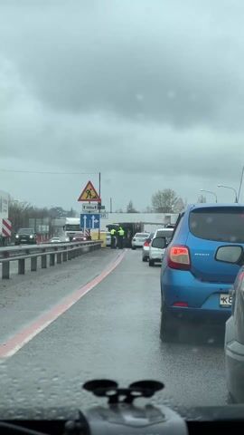 The video shows a traffic jam on a wet road under a cloudy sky. A truck is lying on its side, blocking part of the road. Two police officers in green vests are standing near the overturned truck, talking. Several cars are stopped in the traffic jam, including a blue Renault in the foreground. Traffic signs indicate road work ahead.