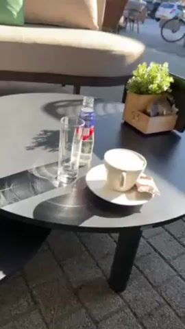The video shows a table outside a cafe.  On the table is a glass, a small bottle of water, a cup of coffee on a saucer with a sugar packet, and a small wooden planter with artificial greenery and a card stand in it.  There is outdoor seating in the background. Bicycles are visible on the sidewalk.  The camera pans around the table.