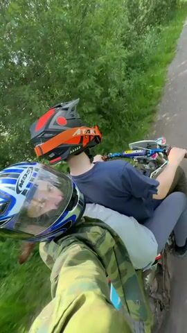 Two individuals are riding a motorcycle on a dirt path surrounded by green foliage under a blue sky with white clouds. The person in front is driving, wearing a black t-shirt, grey pants, and an orange and black full-face helmet. The passenger in the back, wearing a camouflage jacket, a white fanny pack, and a blue and black full-face helmet, is recording the scene from a first-person perspective, looking forward and slightly to the right.