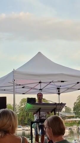 The video shows a man standing behind a keyboard on a stage, playing music. He is wearing a white t-shirt and shorts, and he has a microphone in front of him. There is a white tent over the stage, and two women are sitting in front of the stage, watching the performance. The background shows a body of water with boats, and trees. The sky is cloudy.