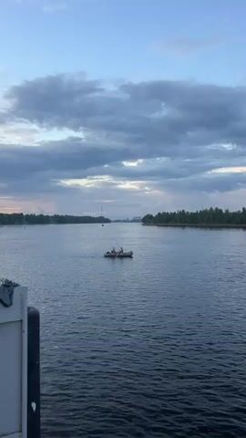 A wide shot shows a calm, dark blue river under a cloudy blue sky with streaks of light orange at the horizon. The left edge of the frame features the corner of a white structure with some blue fabric draped over it. In the middle of the river, a small, dark inflatable boat with two occupants is visible. The boat is pointed towards the left. The far bank of the river is lined with a dense forest. The overall scene is peaceful and serene.