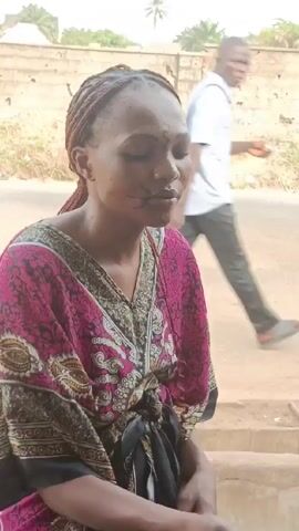 A woman wearing a pink and black patterned robe and braided hair is inspecting a black sandal. She has what appear to be facial markings. The background includes a wall, a street, and some greenery. A person walks by in the background.