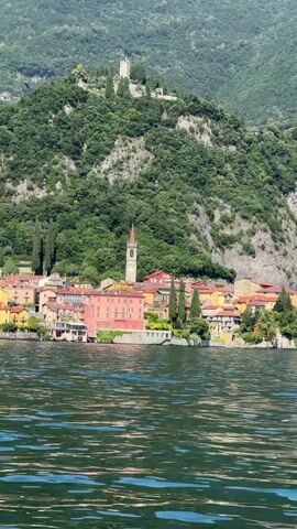 The video shows a waterfront village nestled at the base of a lush, tree-covered mountain. The village has colorful buildings with red-tiled roofs, and a prominent church with a tall, slender steeple. Above the village, a castle or fortress can be seen on the mountaintop. The foreground is dominated by the rippling, dark green water of a lake or sea, with small whitecaps indicating a gentle breeze. The sky is a pale, hazy blue. The camera appears to be moving slowly, possibly from a boat on the water, offering a scenic view of the village and its mountainous backdrop. The overall impression is one of a picturesque, tranquil European landscape.