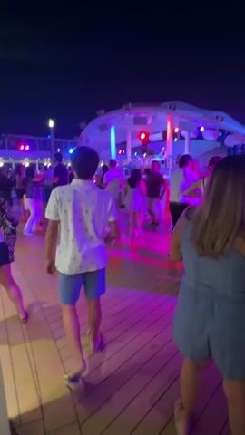 A group of people are dancing on a ship deck at night. The deck is made of wood and is illuminated by purple and pink lights. There is a stage in the background with a band playing. Some people are dancing in pairs, while others are dancing alone. The atmosphere is festive and lively.