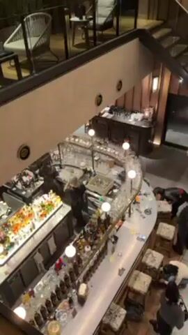 The video shows a bird's-eye view of an upscale bar and lounge area. The bar is a light-colored, curved countertop with numerous bottles and glassware displayed. Two bartenders are working behind the bar, one near a cash register and the other preparing drinks. Several patrons are sitting on high stools at the bar, some looking at their phones or leaning against the bar. A staircase with glass railings leads to an upper level where some wicker chairs and small tables are visible. The walls are decorated with vertical stripes and sconce lighting. The music playing in the background is upbeat.