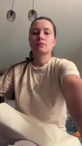 The video shows a woman sitting cross-legged, looking at the camera. She's wearing a beige t-shirt and white pants. In the background, there are two hanging lamps and a partial view of a wall with some decorations. She then picks up a white, drawstring bag and reaches inside to pull out something.