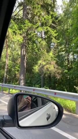 The video shows a view from the passenger side of a car traveling on a road through a forest. The side mirror is visible in the foreground, reflecting a person holding a phone. Tall trees with green foliage line both sides of the road, with a guardrail separating the road from the trees. The road has white markings and other cars are visible in the distance. The sky is blue with some clouds. The overall impression is of a scenic drive through a wooded area on a sunny day.