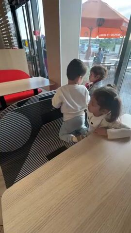 The video shows three young children sitting at a booth in a fast-food restaurant. Two girls and one boy are visible. The girl in the foreground is wearing a purple coat with a white trim and a pink tulle skirt. She is looking to the left. The boy is wearing a white sweater and is looking to the right. Another girl, partially obscured, is sitting between the boy and the girl in the purple coat, and she is also looking to the right. The booth has a black and white striped divider. In the background, through a large window, an outdoor seating area with tables and umbrellas is visible. A woman in a yellow jacket and another child in a pink outfit are walking inside the restaurant. The restaurant interior features red booth seating and wooden tables. A poster advertising "Sauce Curry" is on the wall. The lighting suggests it is daytime.