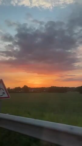 The video shows a sunset over a green field, seen from what appears to be a moving vehicle. The sky has colors ranging from blue to orange, with gray clouds scattered around. There is a line of trees and some buildings in the distance. A white guardrail is visible at the bottom of the frame, indicating the location of the shot. Two people can be heard speaking French in the background.