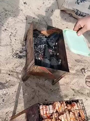 A hand is holding a light green plastic lid over a metal barbecue grill filled with charcoal. Below the grill, another grill holds rows of marinated meat. The scene is set on a sandy surface.